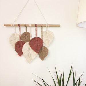 Peut inclure: Une tenture murale en macramé avec sept motifs en forme de feuilles dans des tons crème, rouille et marron. Les feuilles sont suspendues à une cheville en bois naturel avec une ficelle crème pour l'accrochage. Décoration bohème.