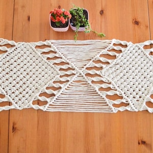 May include: A white macrame table runner with a diamond pattern. The runner is on a wooden table with two small potted plants.