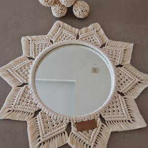 Op de afbeelding: Een witte macram&eacute; spiegel met een stervormig ontwerp en een ronde spiegel in het midden. De spiegel is omgeven door een franje van wit garen.