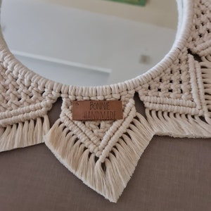 Op de afbeelding: Een witte macram&eacute; spiegel met een bruin label waarop "BONNIE HANDMADE" staat.