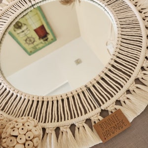 May include: A round macrame mirror with a white cotton cord and fringe. The mirror is reflecting a picture of a lighthouse. The mirror is hanging on a wall with a brown texture. A tag with the text "BONNIE HANDMADE" is attached to the bottom of the mirror.
