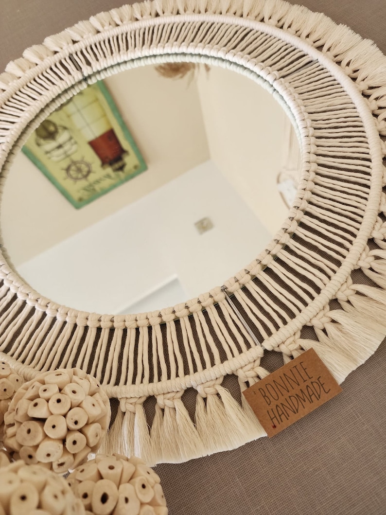 May include: A round macrame mirror with a white cotton cord and fringe. The mirror is reflecting a picture of a lighthouse. The mirror is hanging on a wall with a brown texture. A tag with the text "BONNIE HANDMADE" is attached to the bottom of the mirror.