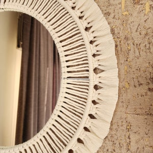 May include: A white macrame mirror with a circular frame and fringe details. The mirror is hanging on a textured wall.