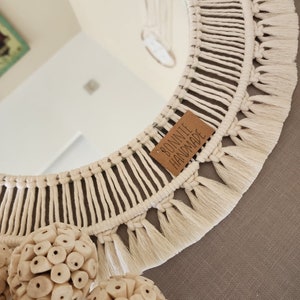 May include: A white macrame mirror with a fringe border and a brown tag that reads "Bonite Handmade". The mirror is hanging on a gray wall with white decorative balls.