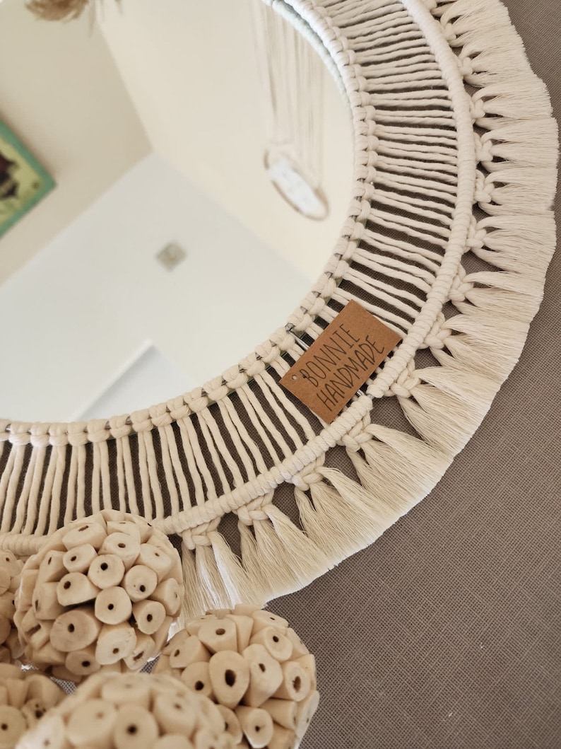 May include: A white macrame mirror with a fringe border and a brown tag that reads "Bonite Handmade". The mirror is hanging on a gray wall with white decorative balls.