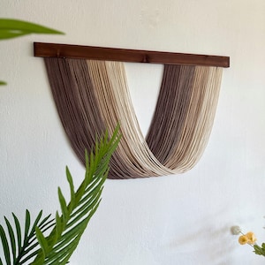 Puede incluir: Un tapiz de macramé con una barra de madera en la parte superior. El colgante presenta hebras verticales en tonos marrones y beige, creando un efecto degradado. La pieza está montada en una pared blanca, con follaje verde en primer plano.