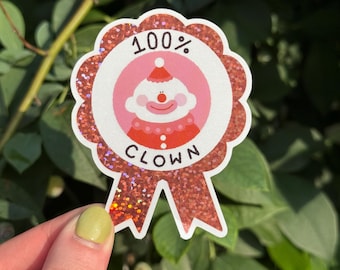 May include: A glittery red sticker with a scalloped edge and a ribbon. The sticker features a pink circle with a cartoon clown and the words "100% CLOWN". The sticker is held against a background of green leaves.