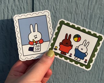 May include: Two stickers featuring cartoon bunnies in a stamp design. One sticker shows a bunny writing, the other shows two bunnies with a ball. The stickers have a white border and are on a blue background.