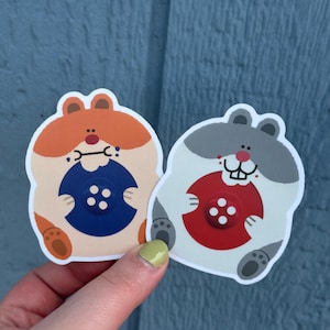 May include: Two die-cut stickers featuring cartoon hamsters. One hamster is orange and cream, holding a blue button. The other is gray and cream, holding a red button. Both stickers have a white border and are held by a hand.