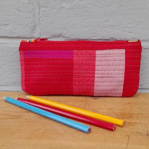 May include: A red quilted pencil case with a zip closure, featuring a patchwork design of various red and pink fabrics. Three coloured pencils, blue, red, and yellow, are lying on a wooden surface in front of the case.