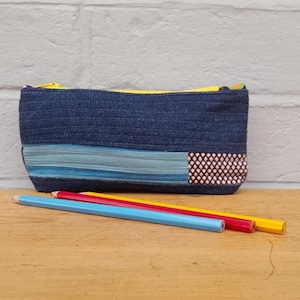 May include: A denim pencil case with a yellow zip, blue and light blue stripes, and a patterned patch. Three coloured pencils in blue, red, and yellow are in front of the case. The case is on a wooden surface.