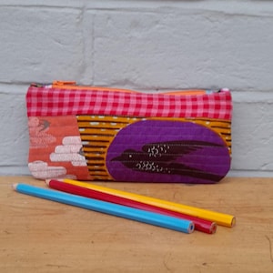 May include: A patterned pencil case with a bird motif, made from orange, yellow, and pink fabrics. The case has a purple oval with a bird design and an orange zip. Three coloured pencils, blue, red, and yellow, are in front.