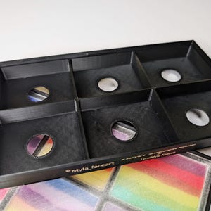 Op de afbeelding: Een zwarte organizer met zes compartimenten en ronde openingen, mogelijk voor make-up of kunstbenodigdheden. De organizer ligt op een kleurrijk, regenboogkleurig oppervlak. De tekst "Myla_faceart" is zichtbaar.