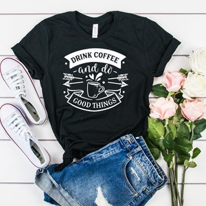 May include: Black t-shirt with white text that reads "Drink coffee and do good things". The text is surrounded by a graphic of a coffee cup, arrows, and a banner. The shirt is laid out on a white wooden surface with a pair of white sneakers and a bouquet of pink roses.