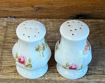 Puede incluir: Un par de saleros y pimenteros de cerámica blanca con un diseño floral. Los saleros tienen una base redonda y una parte superior ligeramente acampanada. El diseño floral presenta rosas y hojas rosas, rojas y verdes.