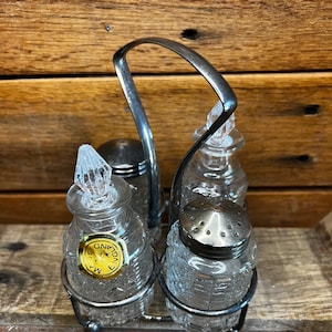 May include: A silver metal salt and pepper shaker holder with four glass shakers. The shakers have a textured design and one shaker has a yellow label that reads "Made in England".