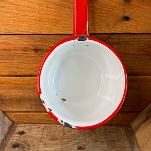 Könnte beinhalten: Rote Emaille-Kasserolle mit weißem Innenraum und rotem Griff. Die Kasserolle hat einen abgenutzten, Vintage-Look.