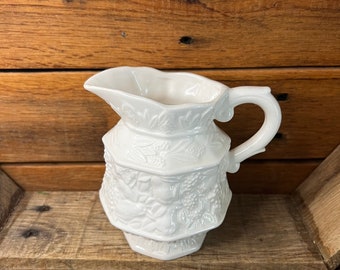 May include: White ceramic pitcher with an embossed floral design. The pitcher has a rounded body and a handle.
