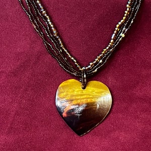 May include: A multi-strand necklace made of brown beads with a large heart-shaped pendant. The pendant is made of a dark brown material with a golden sheen.