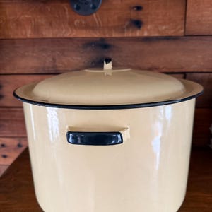 Puede incluir: Una olla de esmalte color crema con tapa a juego. La olla tiene un borde negro y asas negras. La olla está sobre una estantería de madera. Es un artículo de cocina vintage.