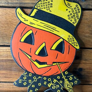May include: Vintage Halloween decoration featuring a smiling orange pumpkin head wearing a yellow and black hat. The pumpkin has black triangle eyes and a yellow mouth with a piece of straw. The pumpkin is on a wooden background.