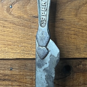 May include: A vintage metal tool with a heart-shaped design and the text "SPERRY" on the handle. The tool has a serrated edge and appears to be used for cutting or prying.