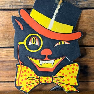 May include: Vintage Halloween cat decoration. The black cat has a top hat, monocle, and bow tie. The hat is yellow and orange, and the bow tie is yellow with red polka dots. The cat is smiling with sharp teeth. Made in the USA.