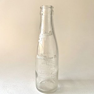 Puede incluir: Una botella de vidrio transparente con una etiqueta blanca que dice "Imitation Grape Flavor" y enumera los ingredientes. La etiqueta tambi&eacute;n incluye el texto "Grapette Bottling Co. Kansas City, Missouri".