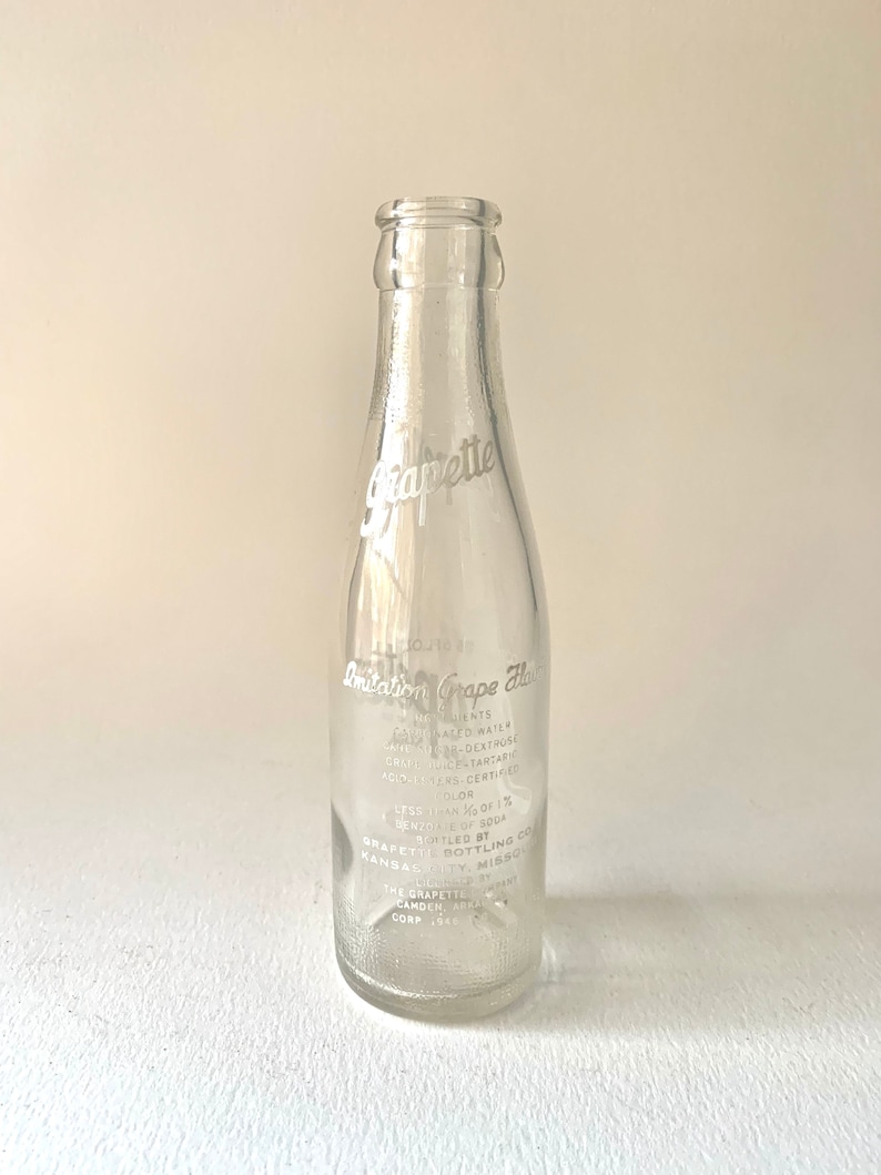 Puede incluir: Una botella de vidrio transparente con una etiqueta blanca que dice "Imitation Grape Flavor" y enumera los ingredientes. La etiqueta tambi&eacute;n incluye el texto "Grapette Bottling Co. Kansas City, Missouri".