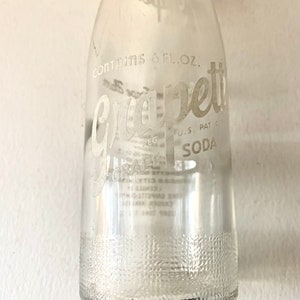 Puede incluir: Una botella de vidrio transparente con una etiqueta blanca que dice "Grape-ette Soda" y "Contains 6 fl. oz."