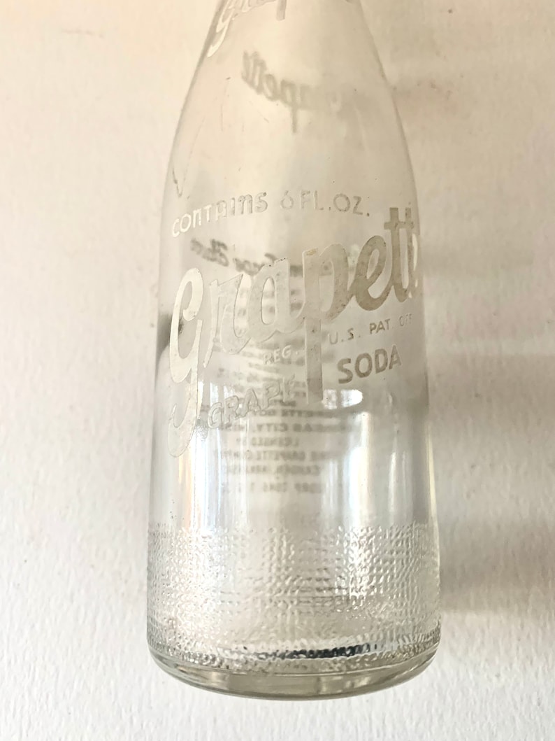 Puede incluir: Una botella de vidrio transparente con una etiqueta blanca que dice "Grape-ette Soda" y "Contains 6 fl. oz."