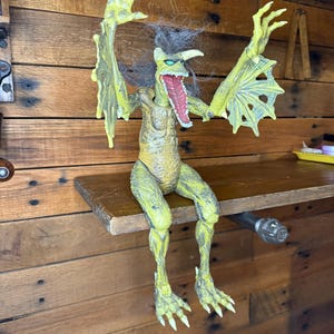 May include: A yellow and green monster action figure with outstretched arms and a wide open mouth. The figure has a detailed, textured design and is posed sitting on a wooden shelf.