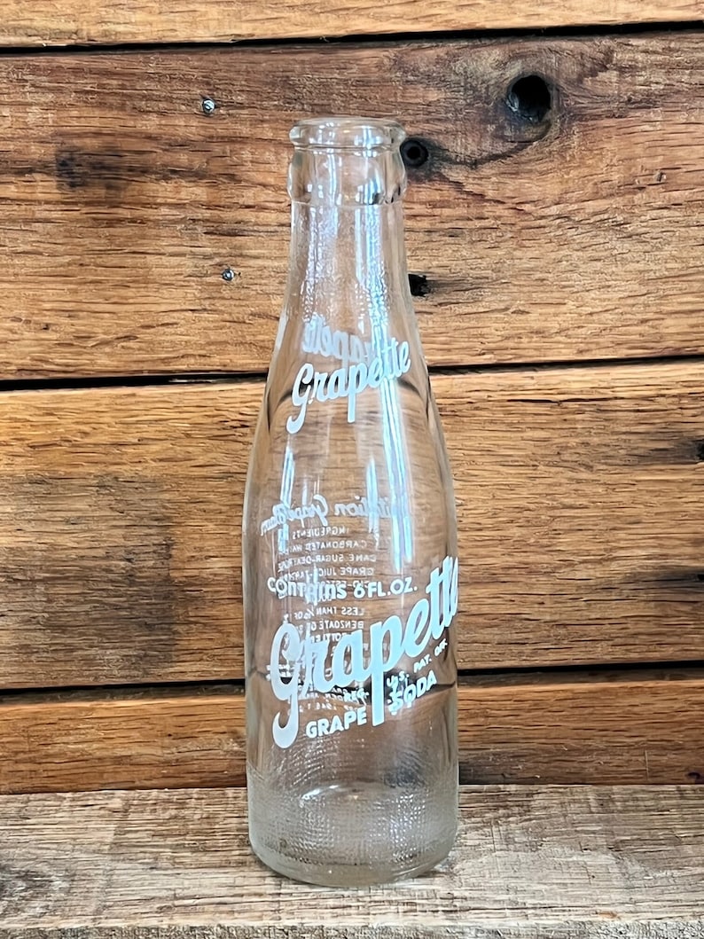 Puede incluir: Una botella de vidrio transparente con una etiqueta blanca que dice "Grape-ette Grape Soda 8 fl. oz. Pat. Off."