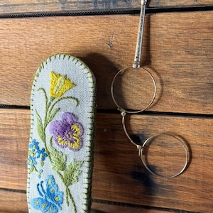 Op de afbeelding: Een antieke bril met gouden montuur en een lange steel, naast een lichtgekleurde etui. De etui is geborduurd met een gele bloem, een paarse en gele viooltje, blauwe bloemen en een blauwe vlinder. De achtergrond is een houten oppervlak.