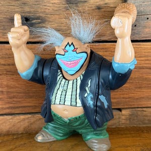 Peut inclure: Figurine de clown avec un visage bleu, des lèvres roses et des cheveux blancs hérissés. Le clown porte une veste noire, une chemise rayée et un pantalon vert. Le clown lève les mains, l'une avec un doigt pointé vers le haut et l'autre tenant un burger.