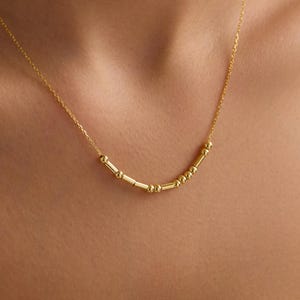 Puede incluir: Un delicado collar de oro con una serie de pequeñas cuentas y barras cilíndricas espaciadas uniformemente. El collar se usa sobre una parte superior de color claro, resaltando su diseño sutil. La joya es un accesorio minimalista.