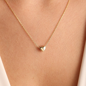 Könnte beinhalten: Eine zarte goldene Halskette mit einem kleinen herzförmigen Anhänger. Der Anhänger hängt an einer feinen Kette und erzeugt ein minimalistisches Design. Die Halskette wird vor einem weichen, neutralen Hintergrund gezeigt.