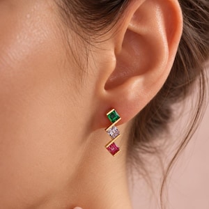 May include: A gold-toned earring featuring three square-cut gemstones arranged in a vertical, staggered zigzag pattern. The top stone is emerald green, the middle stone is light purple, and the bottom stone is a vibrant magenta pink.