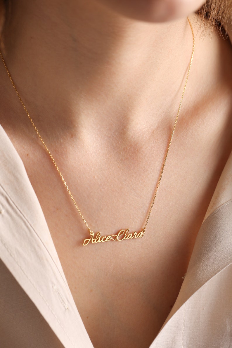 May include: Gold necklace with a delicate chain and a personalized nameplate pendant that reads "Alice Clara" with a heart symbol between the names.