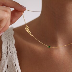 K&ouml;nnte beinhalten: Eine zarte goldfarbene Halskette mit einem fl&uuml;gelf&ouml;rmigen Anh&auml;nger und einem kleinen gr&uuml;nen Edelstein. Die Halskette wird vor einem neutralen Hintergrund pr&auml;sentiert, der ihr elegantes Design hervorhebt. Die Kette ist fein und der Fl&uuml;gelanh&auml;nger detailliert.