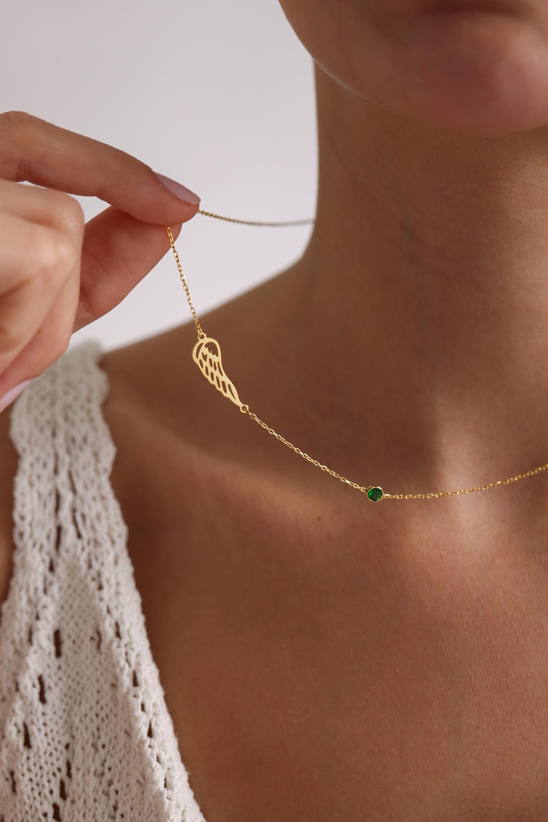 K&ouml;nnte beinhalten: Eine zarte goldfarbene Halskette mit einem fl&uuml;gelf&ouml;rmigen Anh&auml;nger und einem kleinen gr&uuml;nen Edelstein. Die Halskette wird vor einem neutralen Hintergrund pr&auml;sentiert, der ihr elegantes Design hervorhebt. Die Kette ist fein und der Fl&uuml;gelanh&auml;nger detailliert.