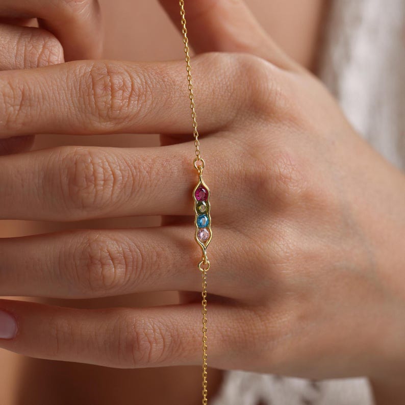 May include: A gold chain bracelet with a pea pod charm featuring five colorful gemstones. The charm is in the shape of a pea pod with five gemstones in a row, representing the peas inside the pod. The gemstones are pink, green, blue, yellow, and red.