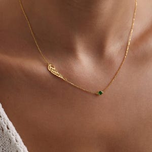 K&ouml;nnte beinhalten: Eine zarte goldene Halskette mit einem fl&uuml;gelf&ouml;rmigen Anh&auml;nger und einem kleinen gr&uuml;nen Edelstein. Die Halskette wird um den Hals getragen und unterstreicht ihr elegantes Design. Der Fl&uuml;gelanh&auml;nger befindet sich links, der Edelstein rechts.