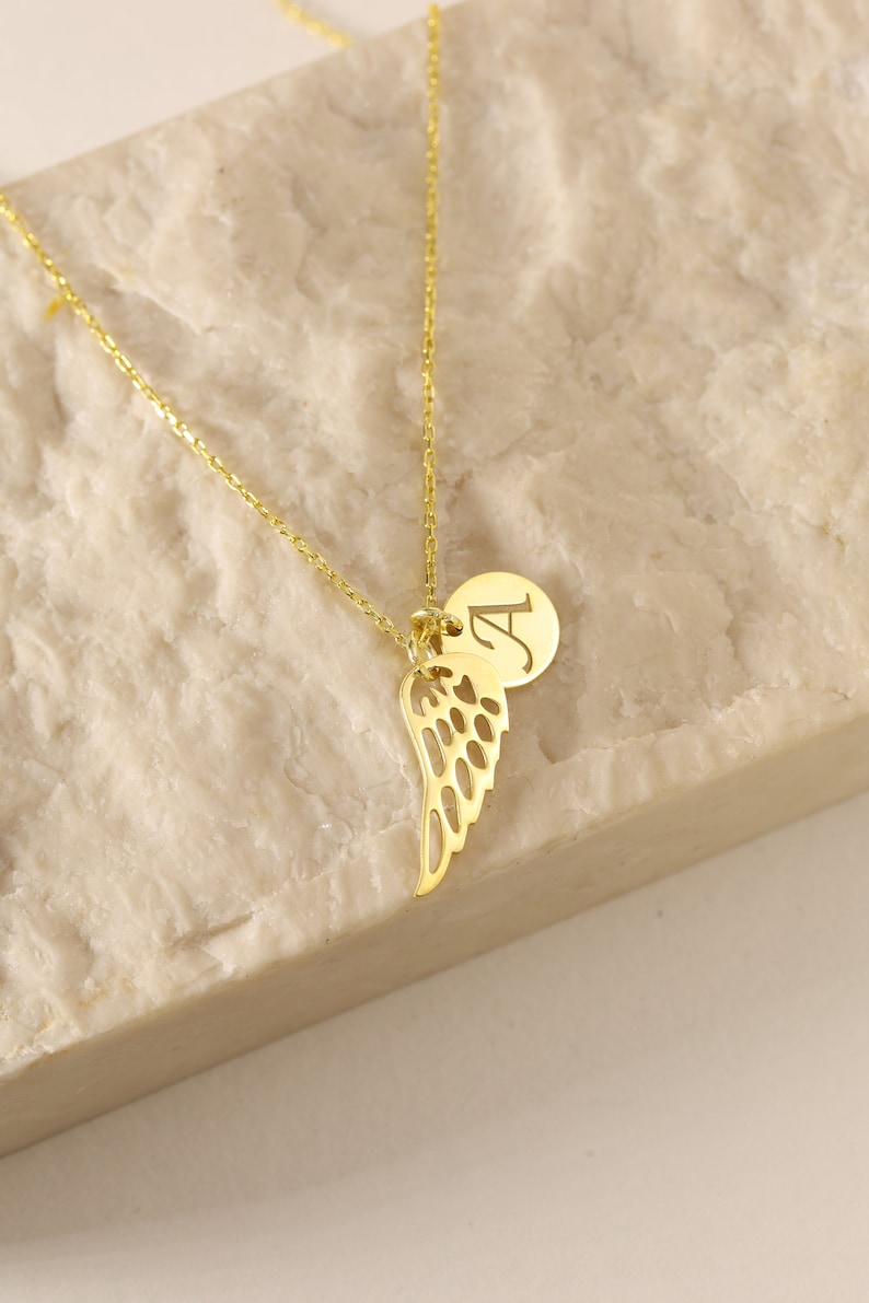 Puede incluir: Un collar de oro con una cadena delicada y un colgante que presenta un ala de &aacute;ngel individual y un peque&ntilde;o dije redondo con la letra "A" grabada en &eacute;l.