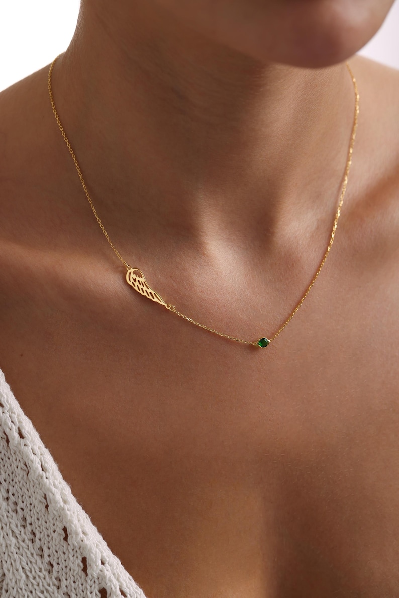 K&ouml;nnte beinhalten: Eine zarte goldene Halskette mit einem fl&uuml;gelf&ouml;rmigen Anh&auml;nger und einem kleinen gr&uuml;nen Edelstein. Die Halskette wird um den Hals getragen und zeigt ihr elegantes Design. Der Fl&uuml;gelanh&auml;nger verleiht dem Schmuck einen Hauch von Laune.