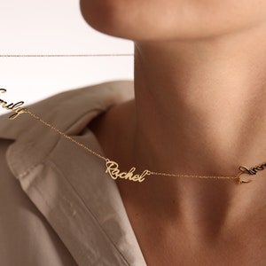 May include: A gold chain necklace with three names on it: Emily, Rachel, and Laura. The necklace is worn by a person with a light brown shirt.
