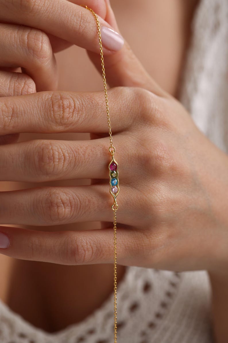 May include: A gold chain bracelet with a pea pod charm featuring five colorful gemstones. The charm is in the shape of a pea pod with five gemstones in a row, each a different color: pink, blue, green, yellow, and red.