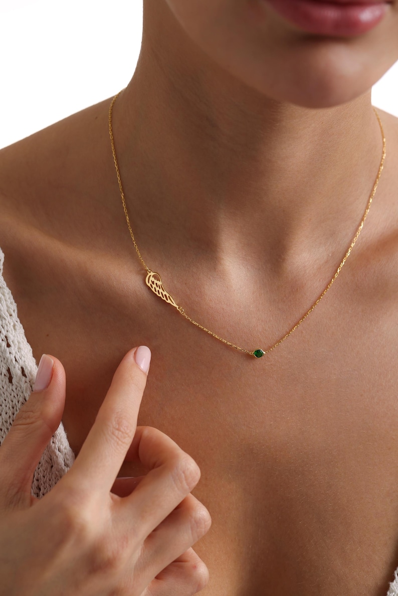K&ouml;nnte beinhalten: Eine zarte goldene Halskette mit einem fl&uuml;gelf&ouml;rmigen Anh&auml;nger und einem kleinen gr&uuml;nen Edelstein. Die Halskette wird am Hals einer Person getragen, wobei sich der Fl&uuml;gelanh&auml;nger links und der Edelstein rechts befindet.