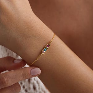 May include: A gold chain bracelet with a pea pod charm featuring five colorful gemstones.