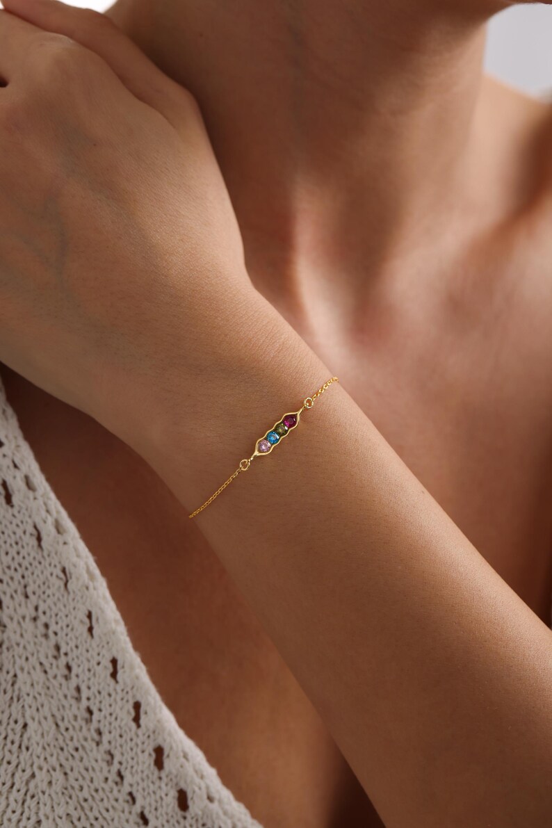 May include: A gold chain bracelet with a small, rectangular charm set with colorful gemstones. The charm is in the shape of a pea pod.